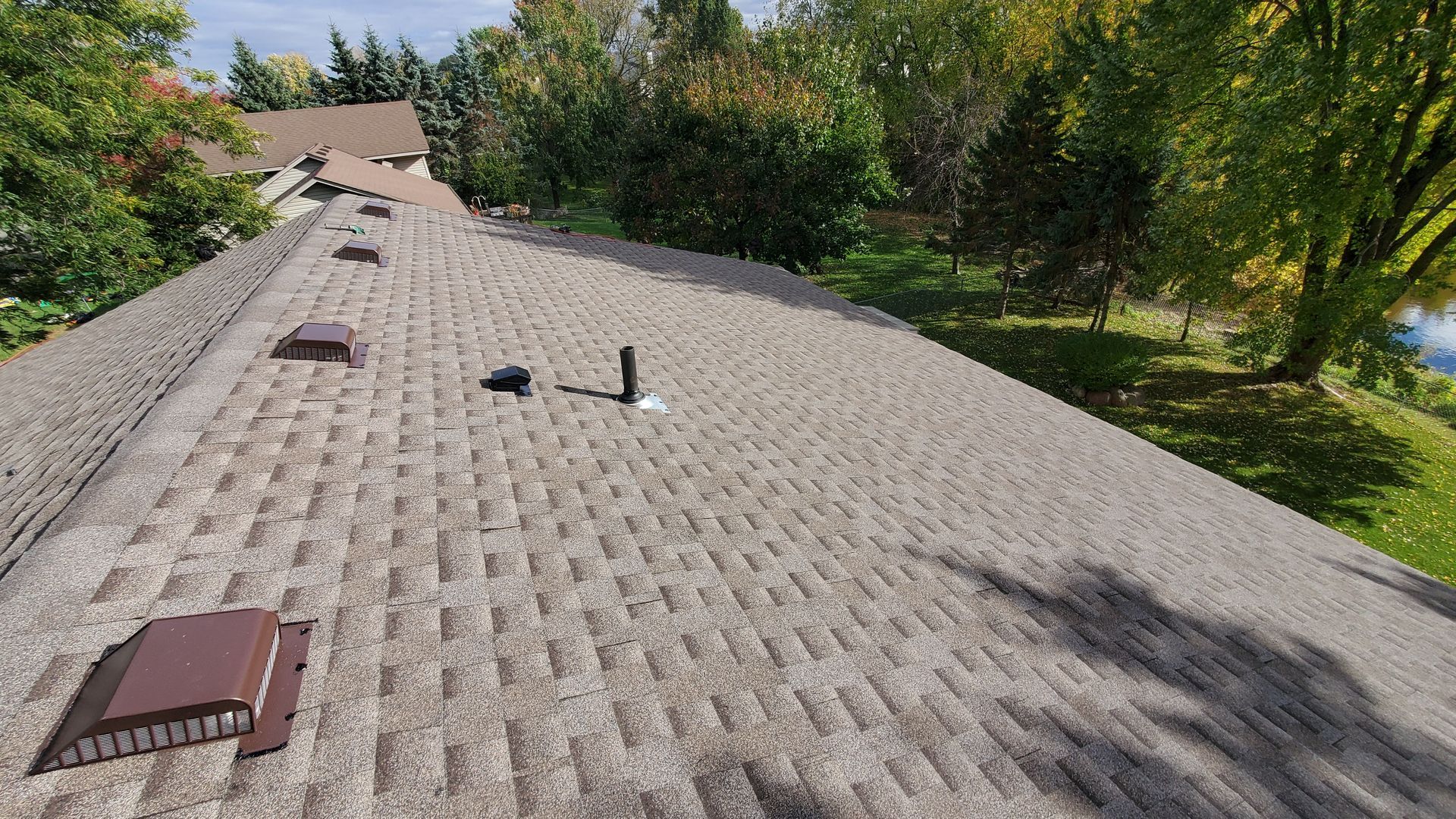 Rooftop covered in gray shingles with vents, surrounded by trees and sunlight