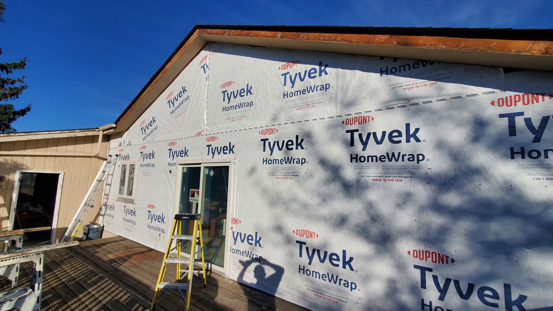 House under construction wrapped in Tyvek, with a ladder and blue sky visible.