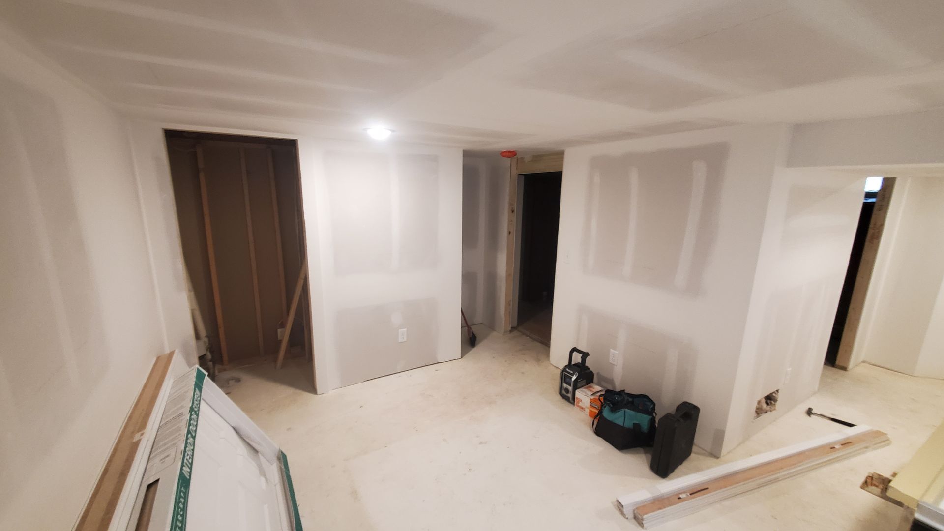 Partially finished room with drywall, a dark door, ceiling light, and tools on the floor.