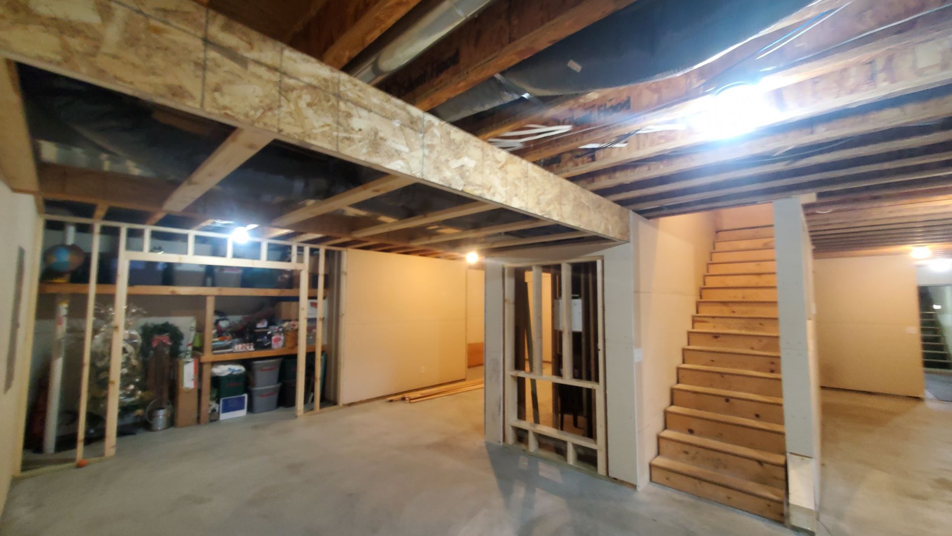 Unfinished basement with exposed beams, concrete floor, storage area, and wooden stairs leading up