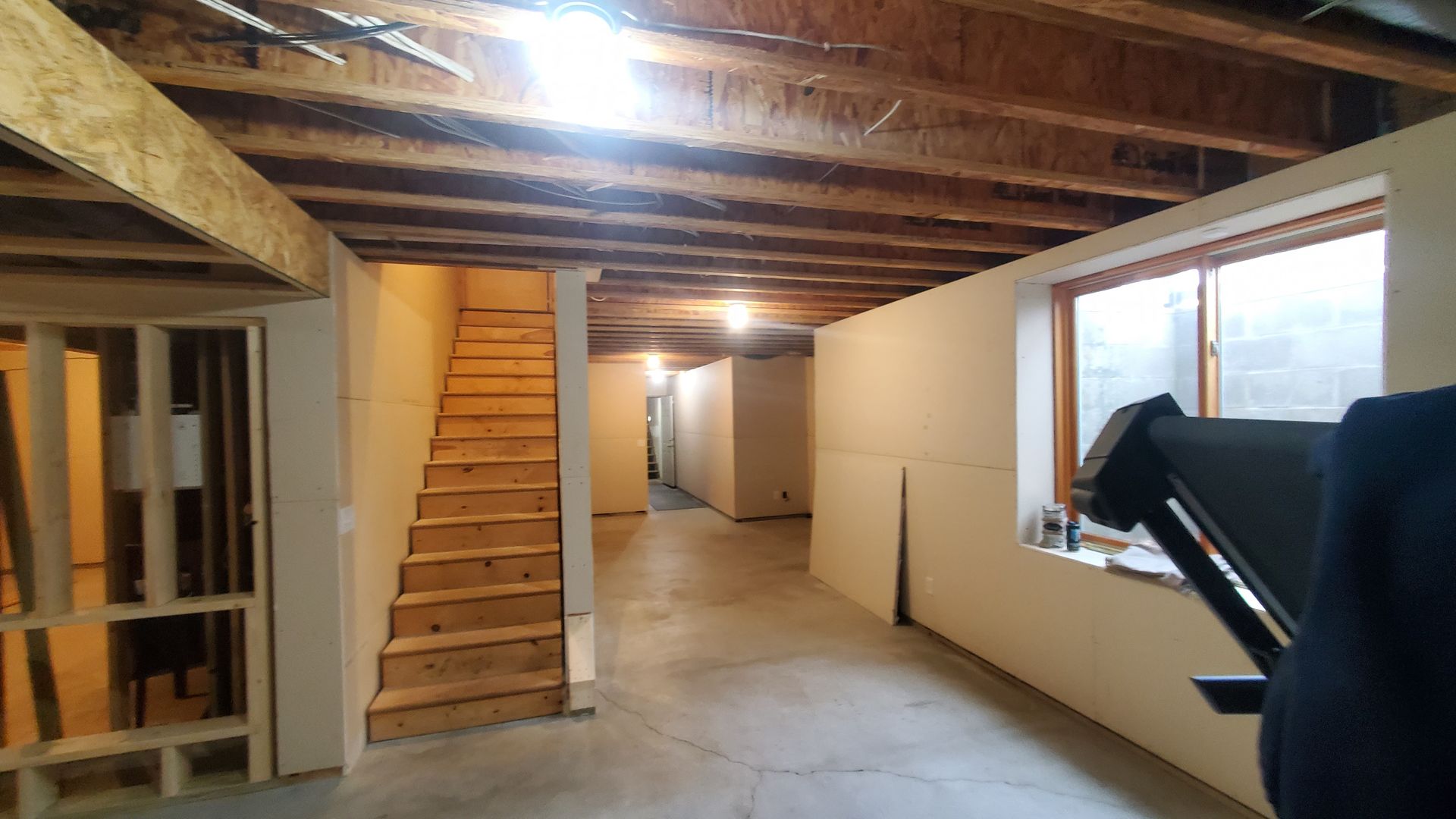 Unfinished basement with exposed beams, stairs, hallway, and a window on the right.