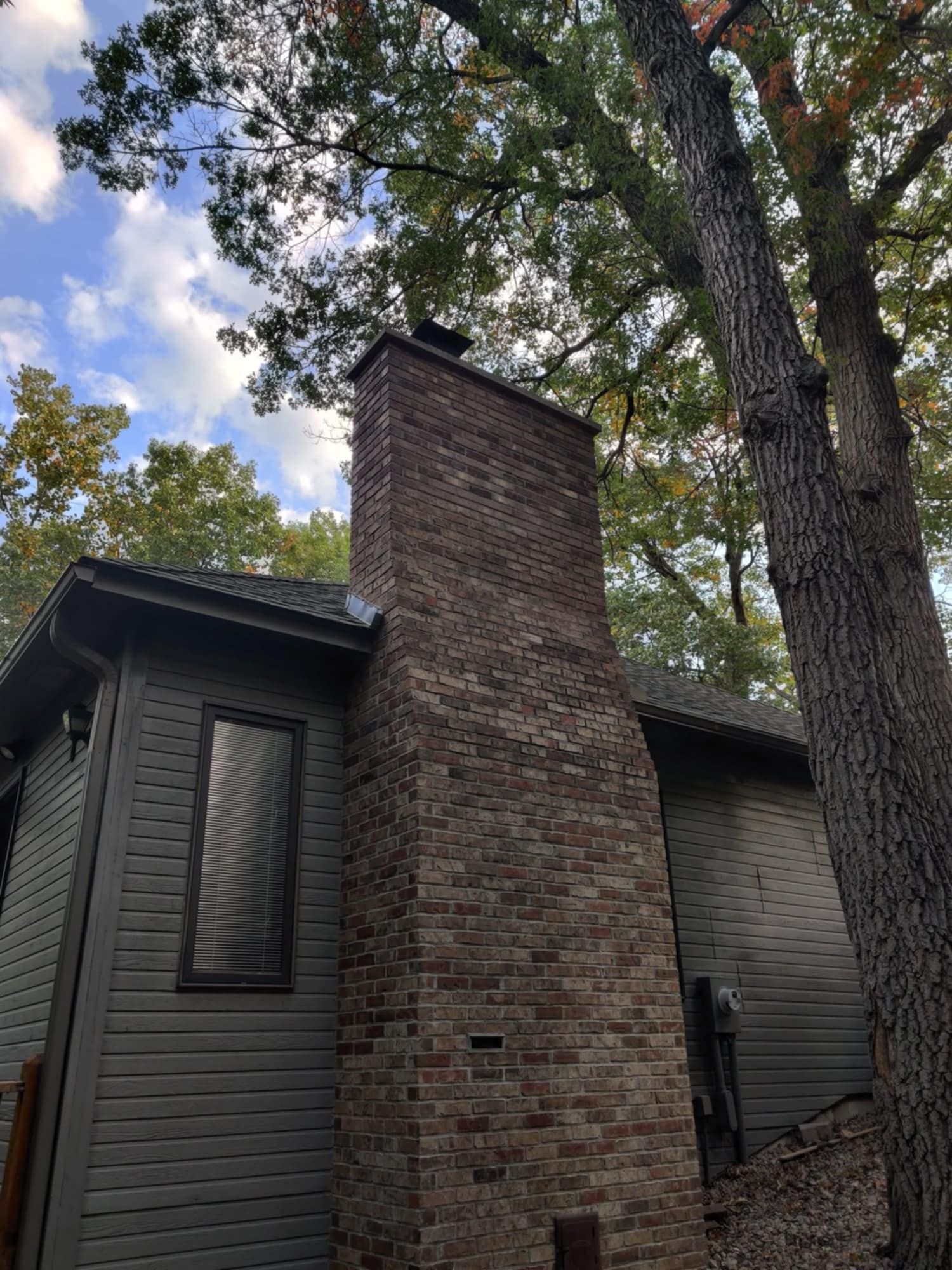 Brick chimney on a dark house, framed by tall trees and a cloudy blue sky.