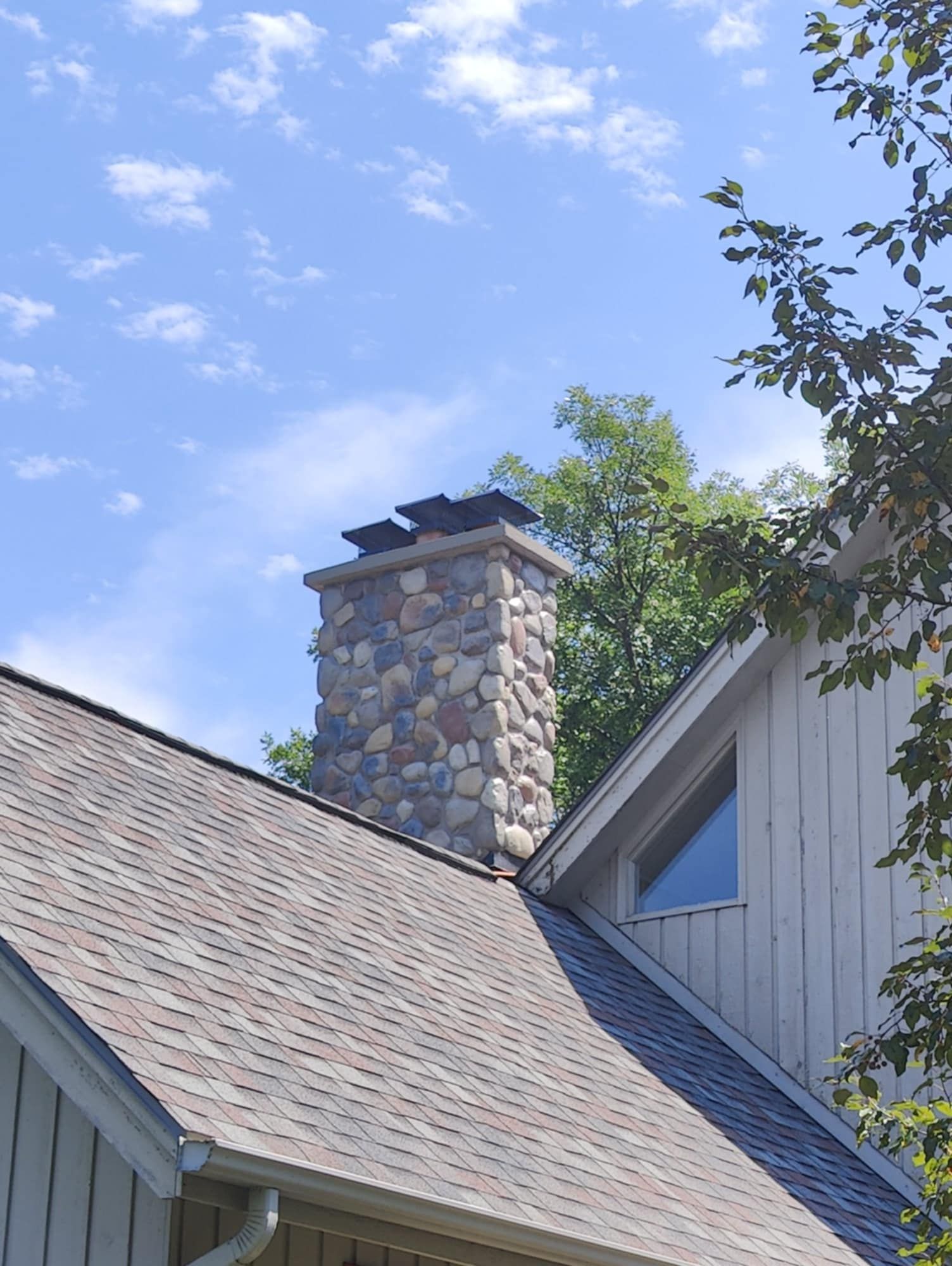 Stone chimney on a house with gray shingled roof under a blue sky and trees