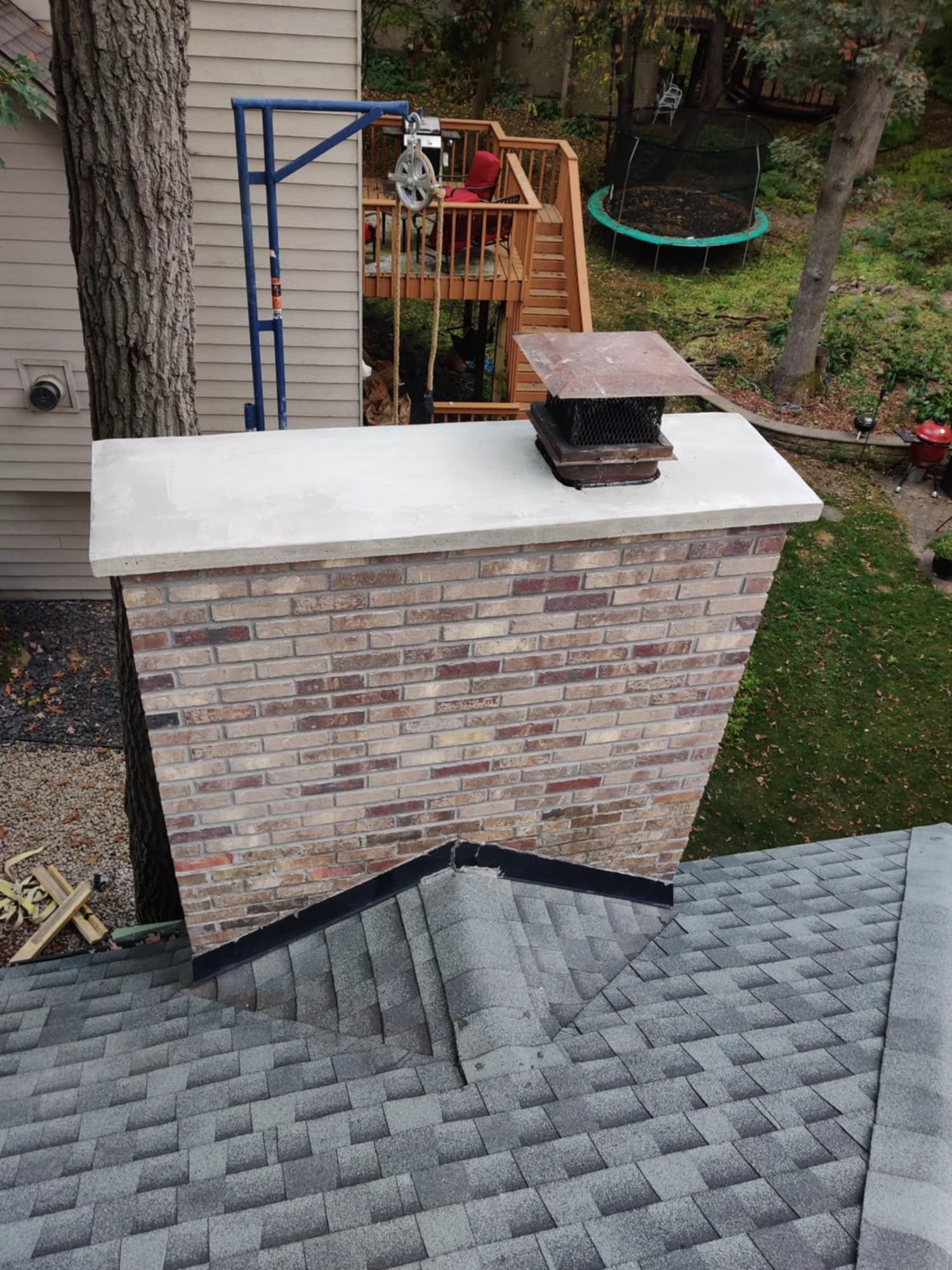 Rooftop chimney with brick surround and metal cap, overlooking a backyard with trees and a trampoline.