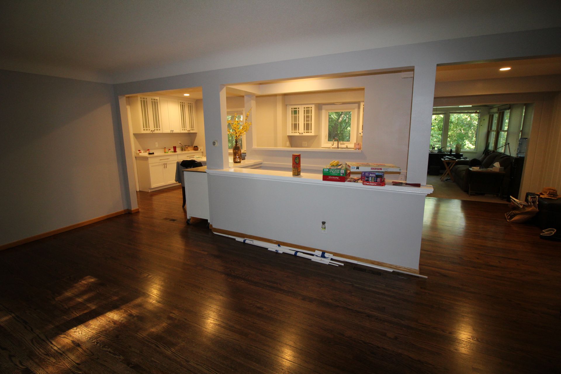 Empty living room with dark wood floors, white half wall, and a bright adjoining kitchen area.