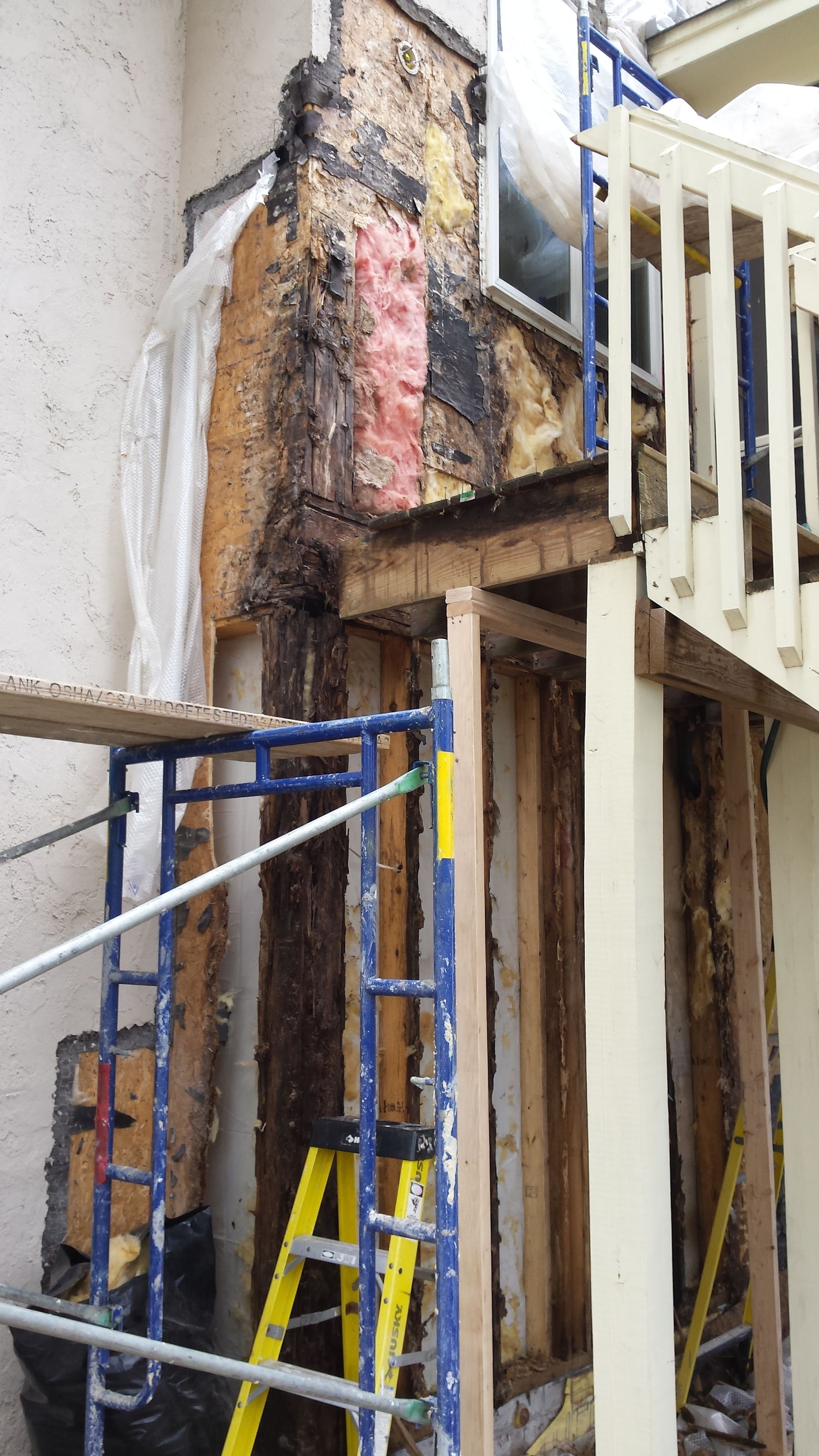 Damaged exterior wall with exposed wood and scaffolding beside a white railing