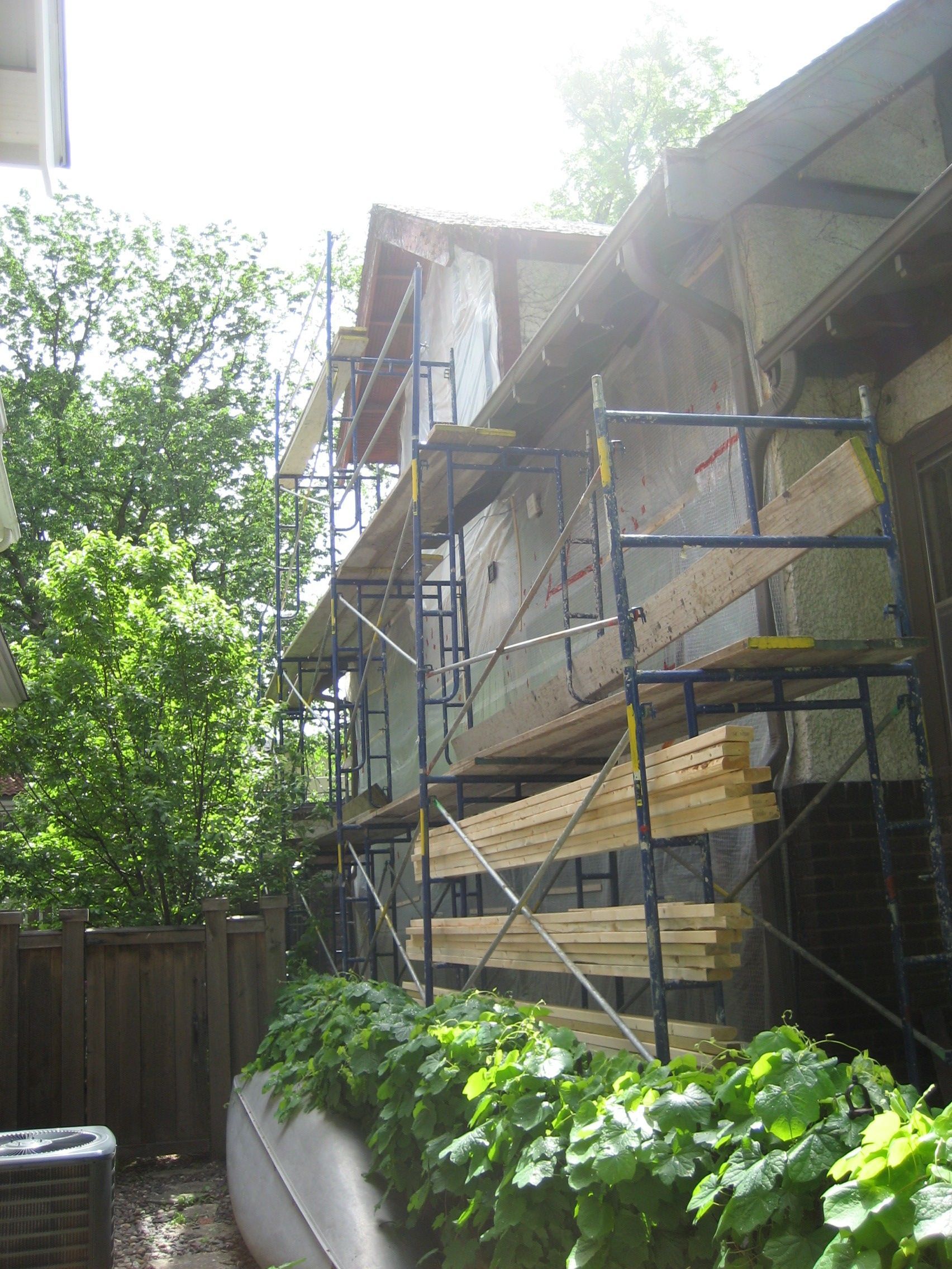 House under construction with blue scaffolding beside a leafy backyard fence