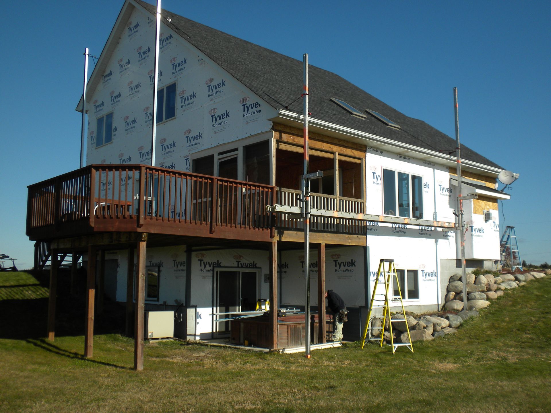 Two-story white house with a wooden deck and balcony on a grassy hillside under blue sky