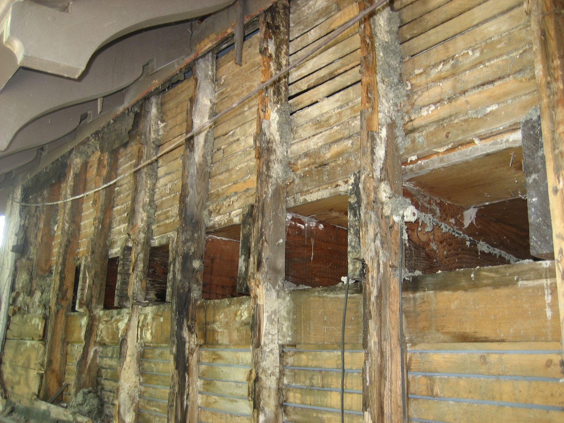 Interior wall with exposed wooden framing and damaged plaster panels, showing heavy water damage.