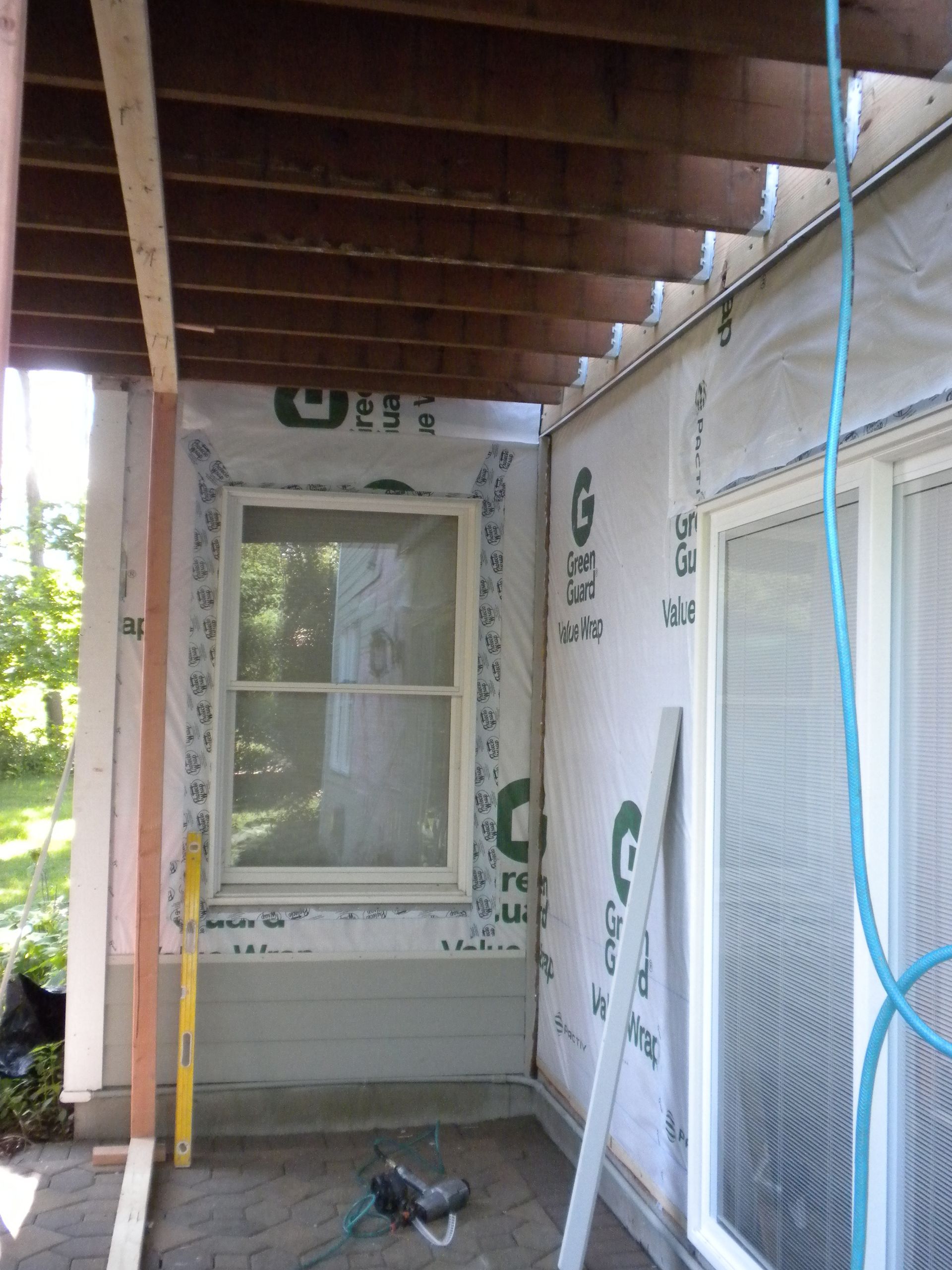 Partially built porch with exposed wood ceiling, window, sliding door, and construction wrap on the walls.