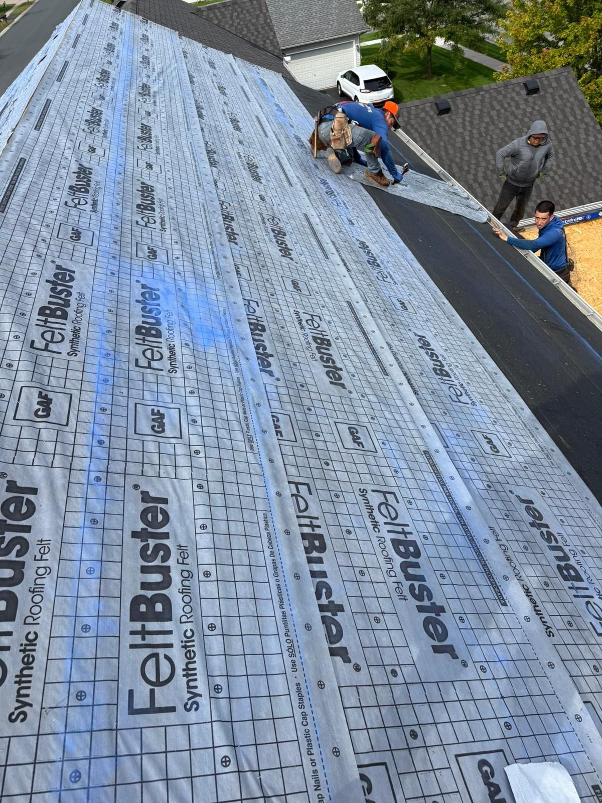 Workers installing roofing underlayment on a sloped roof, with blue and gray synthetic sheets visible.