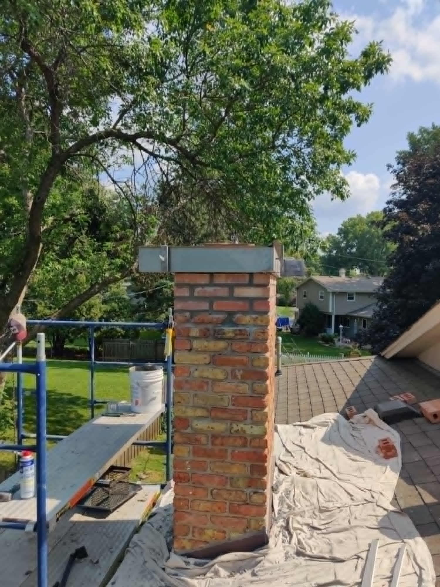 Brick chimney under construction on a rooftop, with a tree and suburban homes in the background.
