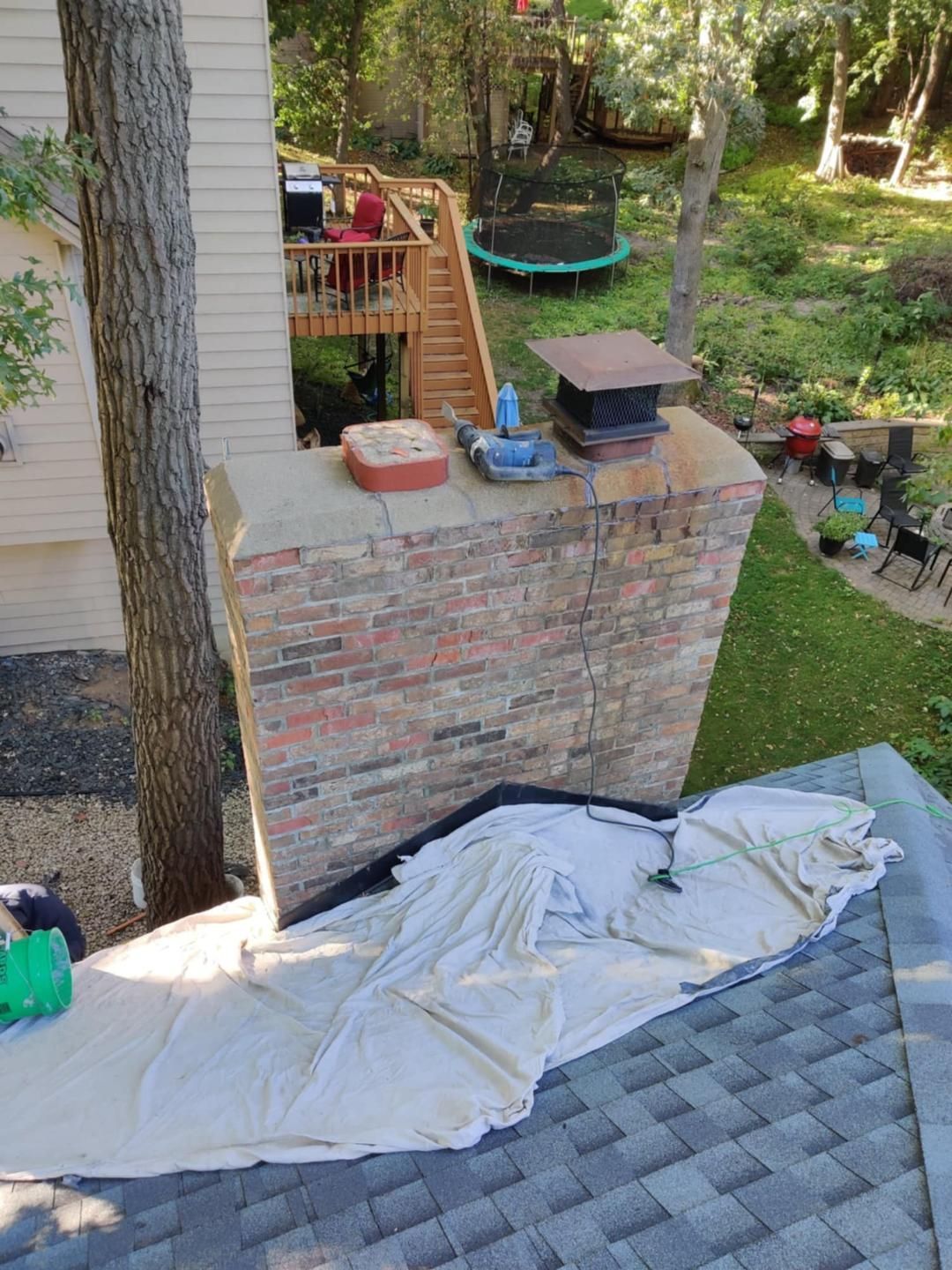 Brick patio wall with potted plants and a trampoline in a leafy backyard; tarp spread across shingles in foreground