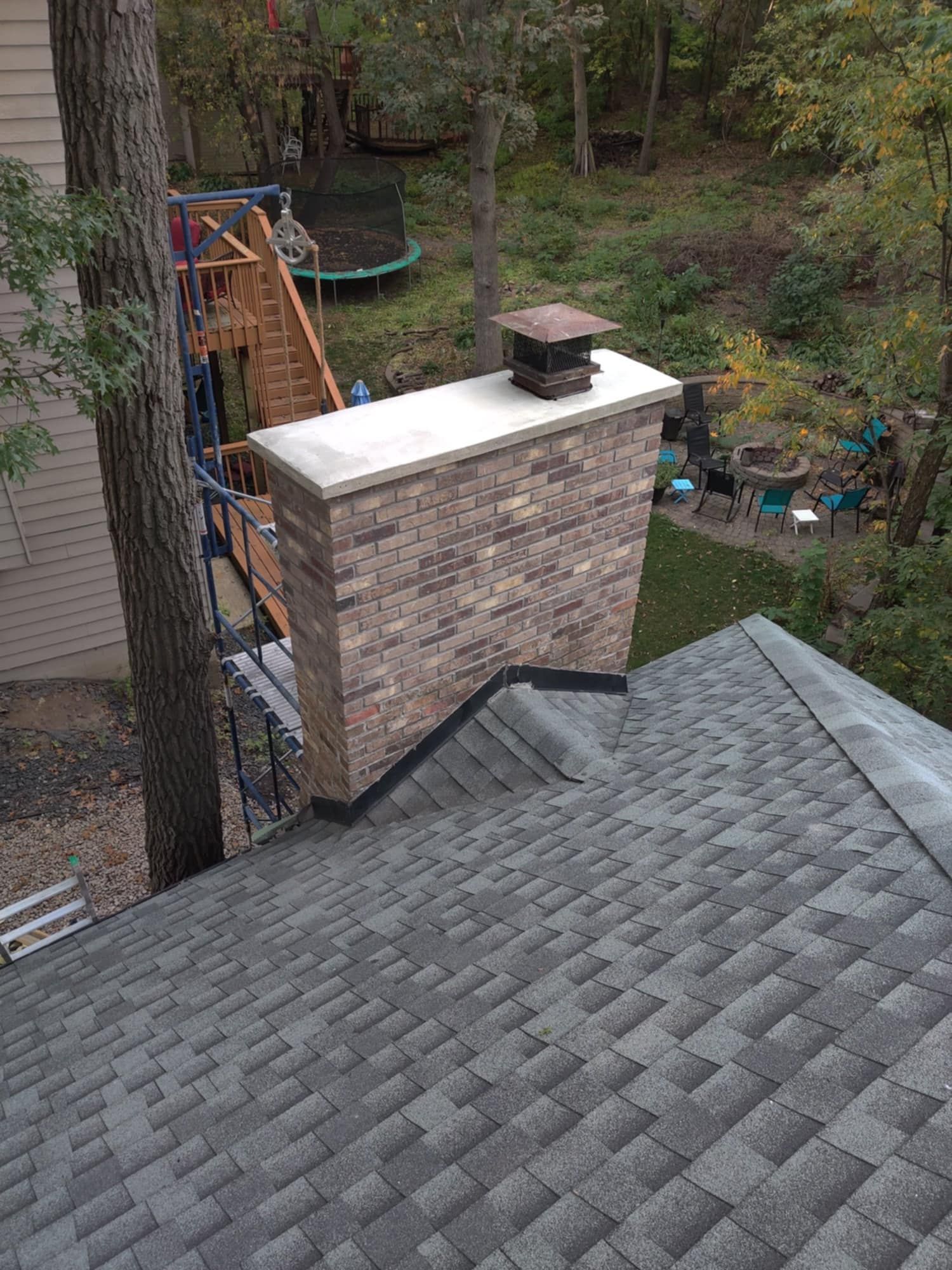 Rooftop view of a brick chimney beside gray shingles, with a wooded backyard and playground in the background