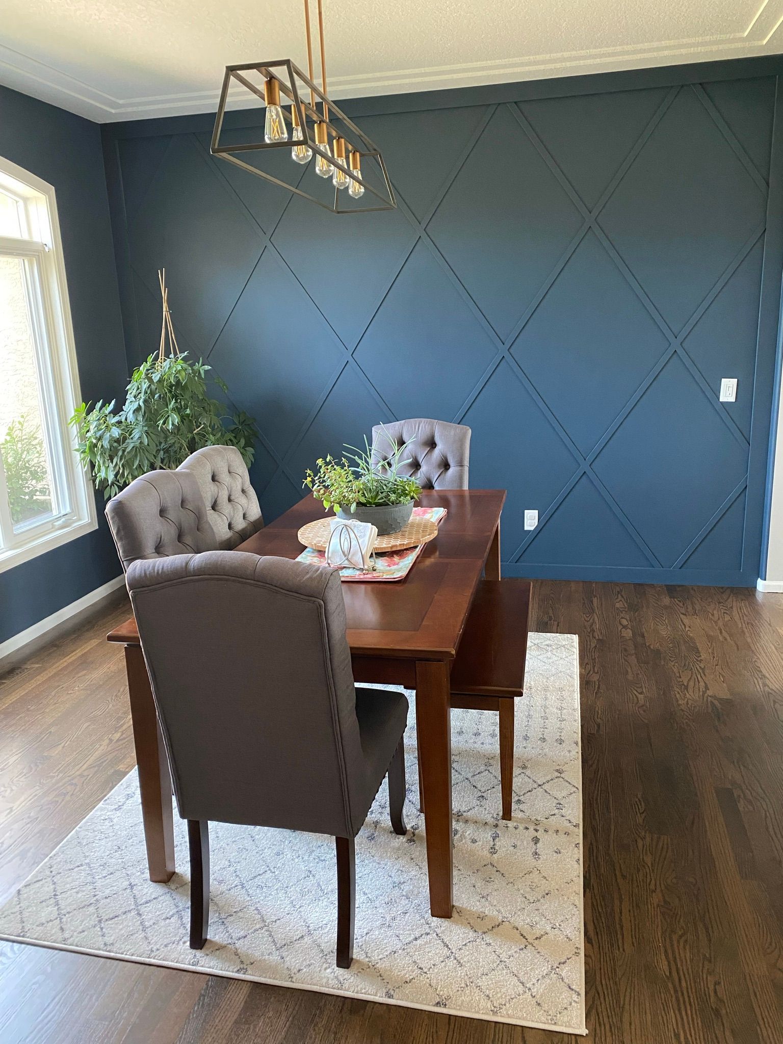 Dining room with dark blue walls, wooden table, tufted chairs, and rug by a bright window.