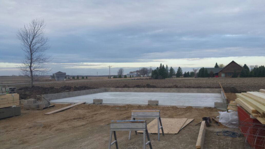 Bare outdoor construction site with concrete foundation, dirt lot, and cloudy sky beyond a rural view