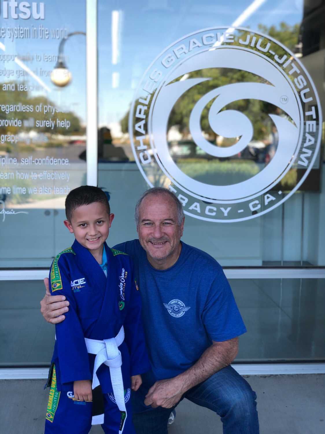 A young boy in a blue gi smiles next to a man in blue; they are in front of a Jiu-Jitsu studio in California.
