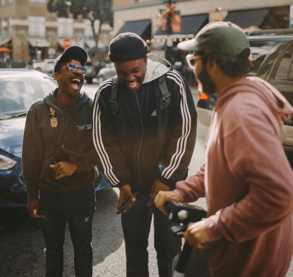 Three people laughing together on a street, with one person in a pink hoodie standing by a bicycle.