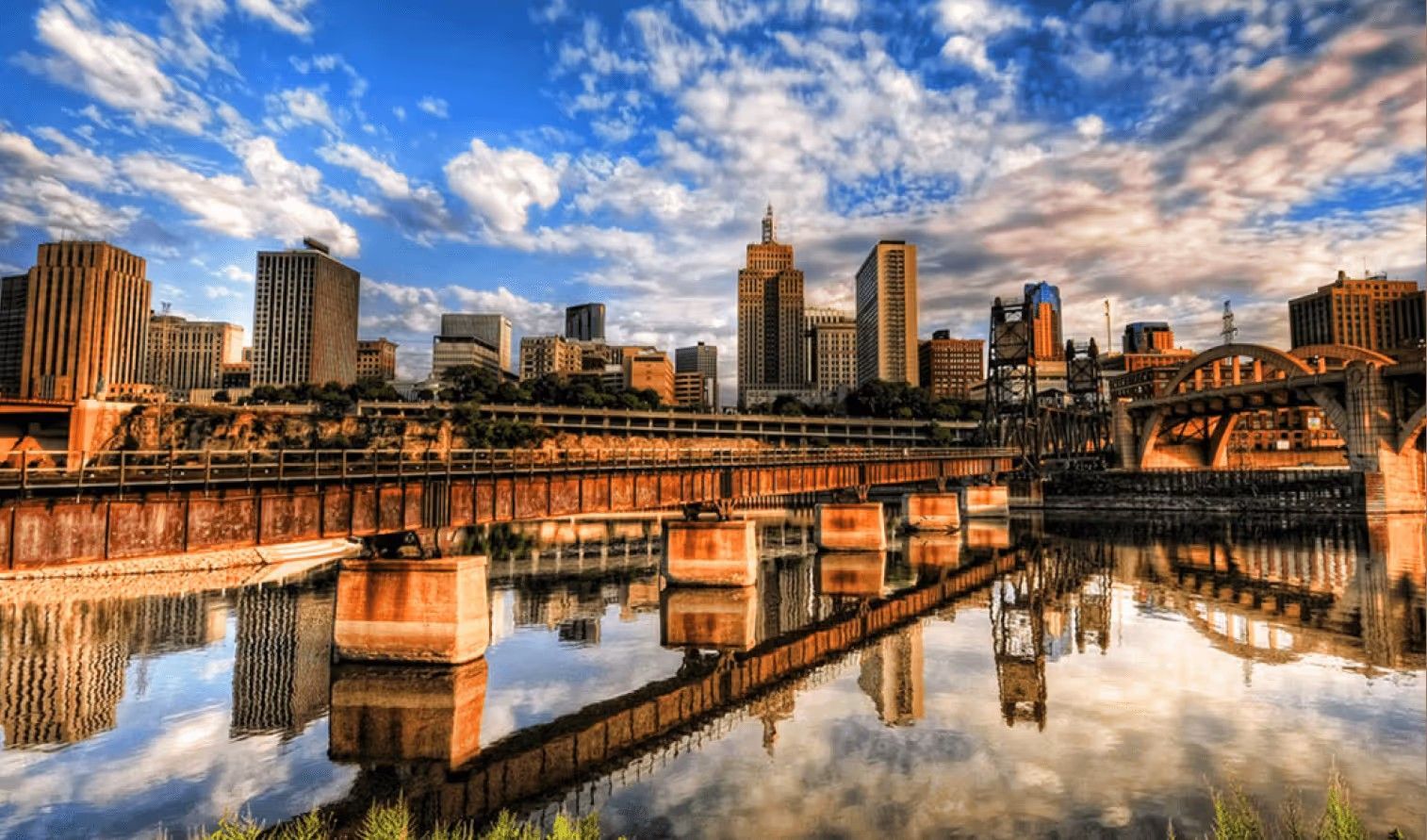 A sunlit cityscape skyline reflected in a calm river, featuring a prominent steel train trestle bridge in the foreground.