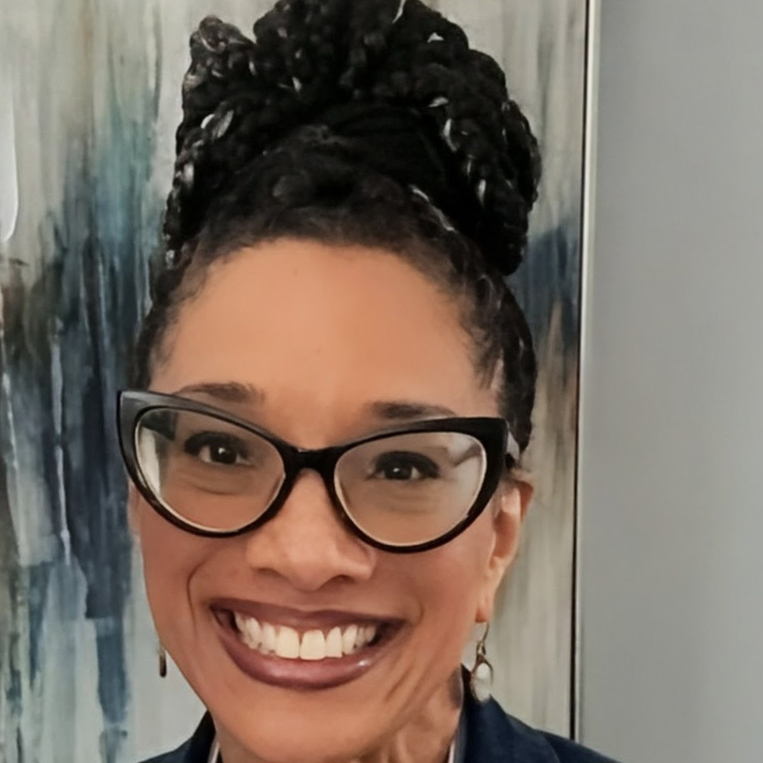 A person with dark curly hair wearing cat-eye glasses, smiling warmly at the camera.