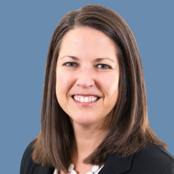 Headshot of a smiling person with shoulder-length brown hair, wearing a dark blazer, against a solid blue background.