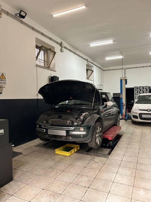 Auto nera con il cofano aperto in un'officina meccanica, sollevata su un ponte, con un'altra auto sullo sfondo.