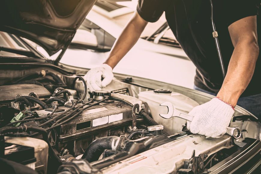 Meccanico con guanti bianchi ispeziona il vano motore con il cofano aperto, utilizzando degli attrezzi in un'officina.