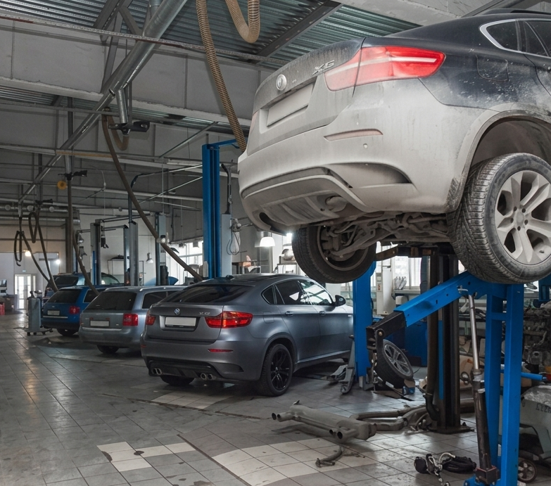 In un'officina meccanica, alcune auto sono sollevate su ponti idraulici, tra cui un SUV argentato in primo piano.