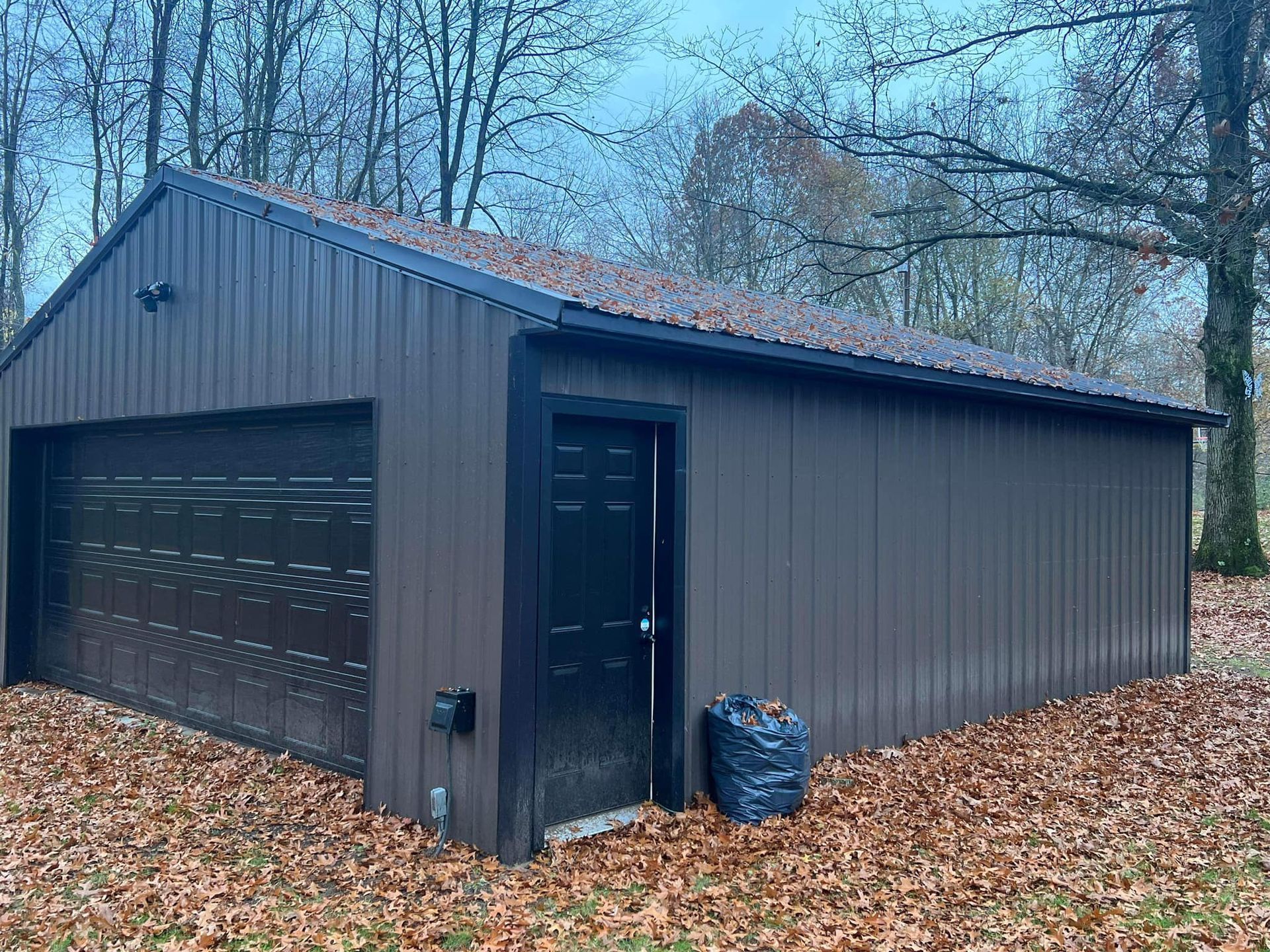 Dark gray metal garage with a side door, set among trees and fallen leaves in a wooded yard.