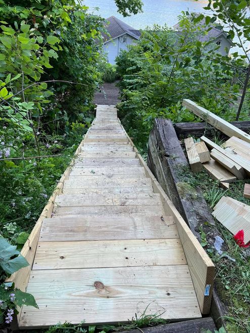 Narrow wooden walkway descending through dense green foliage toward a house, with construction debris on the right