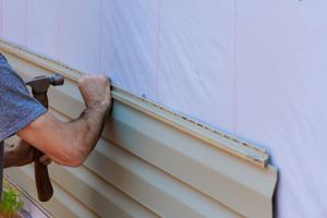 Person installing beige vinyl siding on a house exterior with a hammer