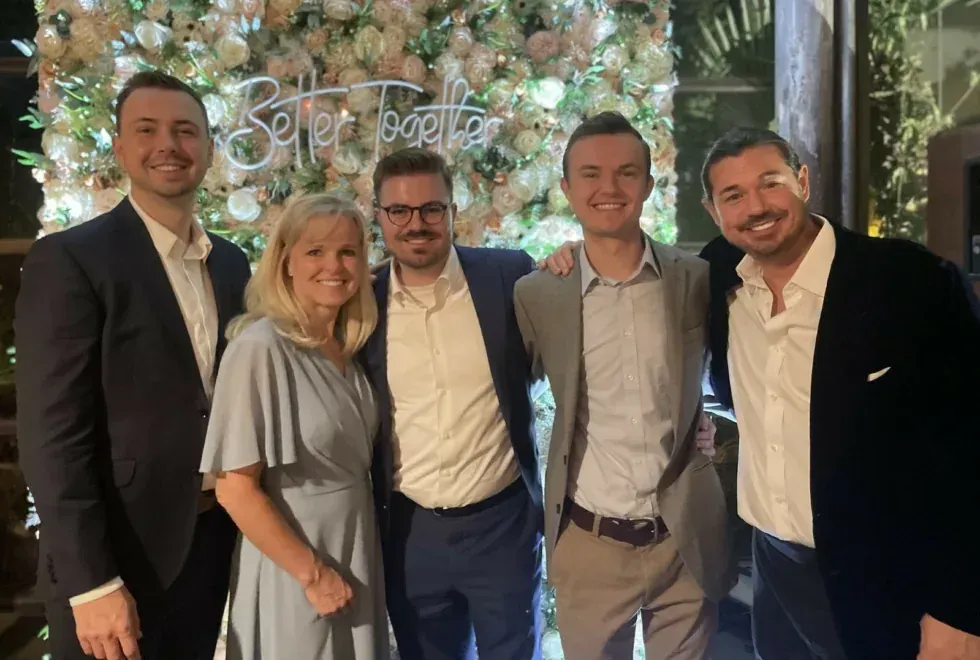 Five people smiling at a formal event with floral lights and a glowing sign in the background
