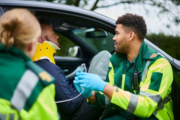 Paramedics assisting a car crash victim beside a damaged vehicle, one wearing a neck brace and oxygen mask.