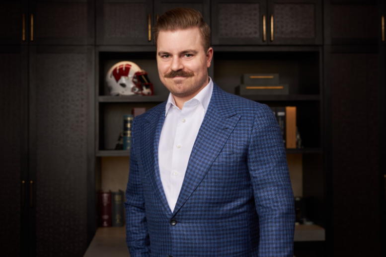 Man in a blue checkered suit stands in front of dark bookshelves, smiling in an office.