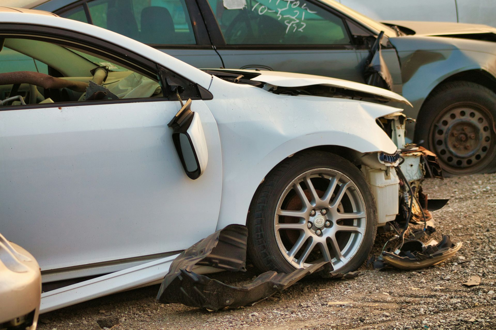 White car with severe front-end crash damage on a roadside, with another damaged vehicle behind it