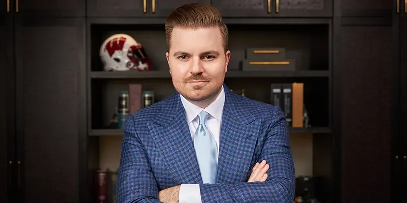 Man in a blue checkered suit stands with arms crossed in an office, shelves behind him.