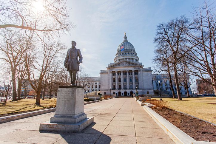 Statue on pedestal before a domed capitol building along a tree-lined walkway