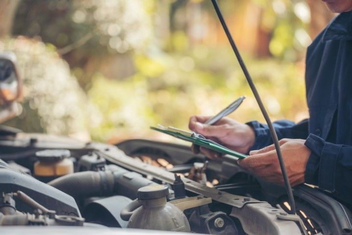 A mechanic in a dark uniform writes on a clipboard while inspecting an open car engine.
