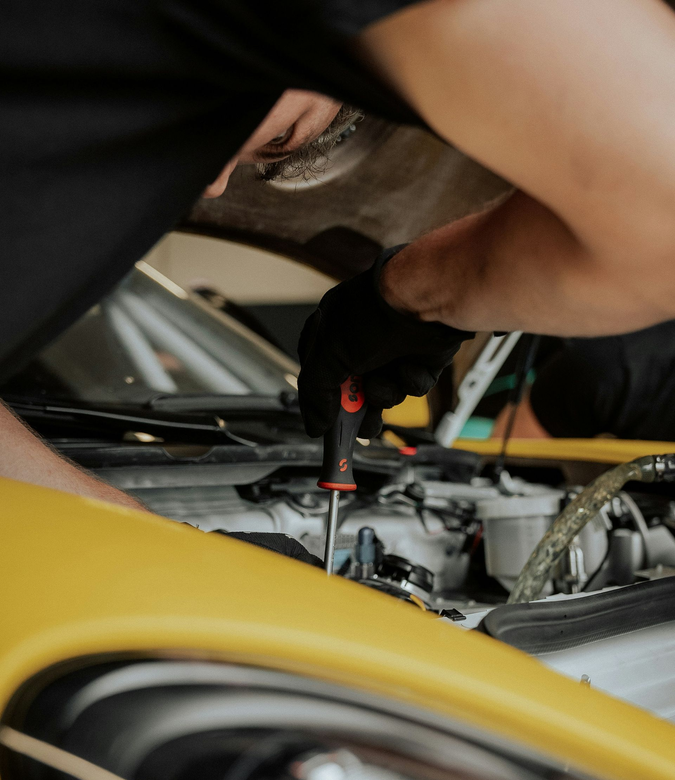 A mechanic in black gloves uses a screwdriver to work on the engine of a yellow car.