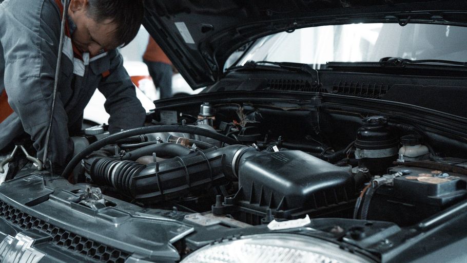 A mechanic in a grey jumpsuit leaning over an open car hood to repair the dark engine components in a workshop.