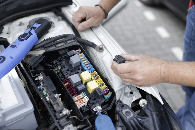 A person works on a car's open fuse box, holding a small electrical component near the brightly colored fuses and relays.