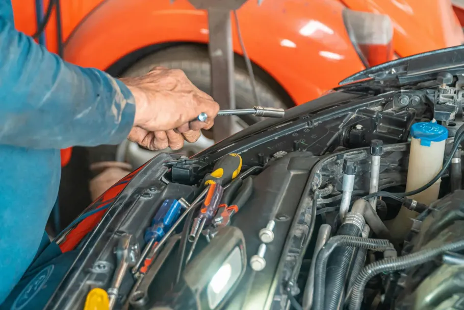 A mechanic uses a tool on an open car engine bay, with other tools and parts laid out nearby.