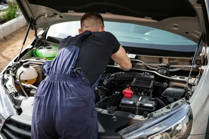 A person wearing dark blue overalls and a black t-shirt leans over the open hood of a car to perform engine repairs.