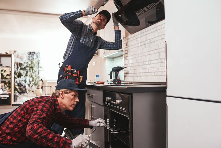 Two people repairing a kitchen oven, one standing and one crouching in a modern kitchen in Chicago