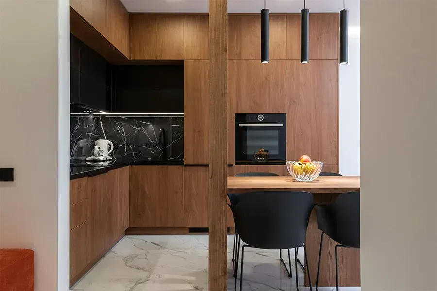 Modern kitchen nook with wood cabinetry, black accents, built-in oven, and a small dining table with chairs.