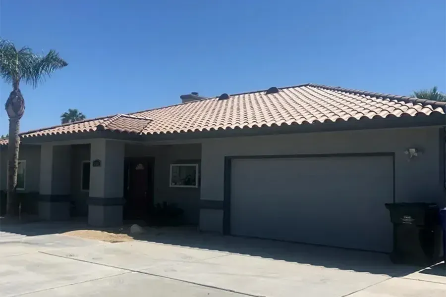 Single-story house with tan tile roof, gray garage door, and palm tree under a clear blue sky