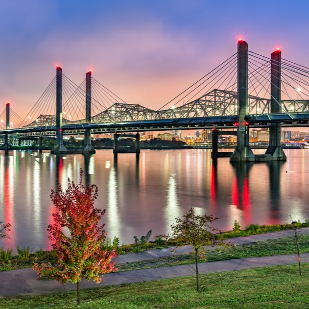 A cable-stayed bridge spans a calm river at sunset, reflecting warm orange and purple light onto the water's surface. Abraham Lincoln Bridge and the John F. Kennedy Memorial Bridge