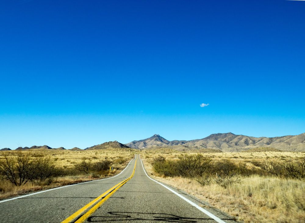 Empty desert highway stretching toward distant mountains under a bright blue sky.