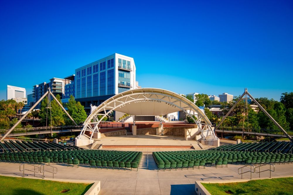 An outdoor amphitheater with rows of green seats faces a stage. A tall building stands in the background under a clear blue sky.