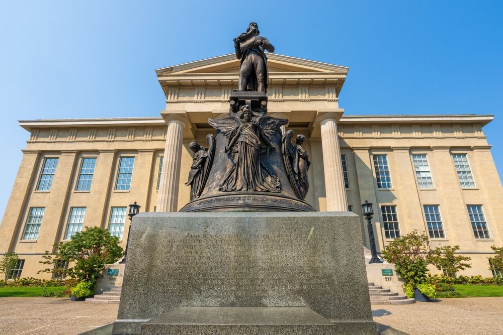 Bronze statue in front of a tan building with columns. Figures surround the statue atop a stone base, under a clear, blue sky. Jefferson Memorial in front of Louisville Metro Hall